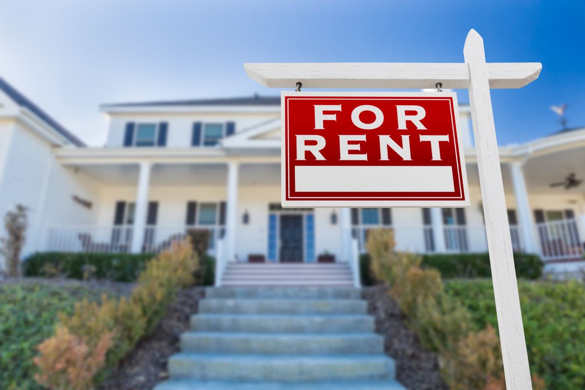 A red "for rent" sign in front of a large white home.