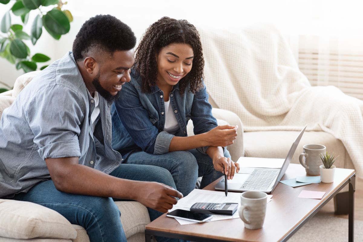 A Black couple sits at their coffee table in front of a laptop and calculator.