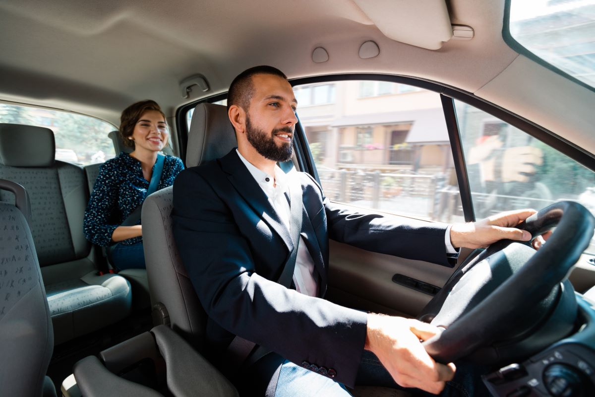 A man drives a car with a female passenger in the back seat.