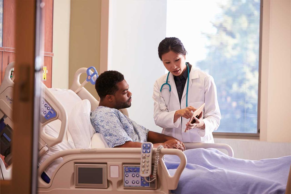 Man sits in hospital bed while doctor shows him something on a tablet.