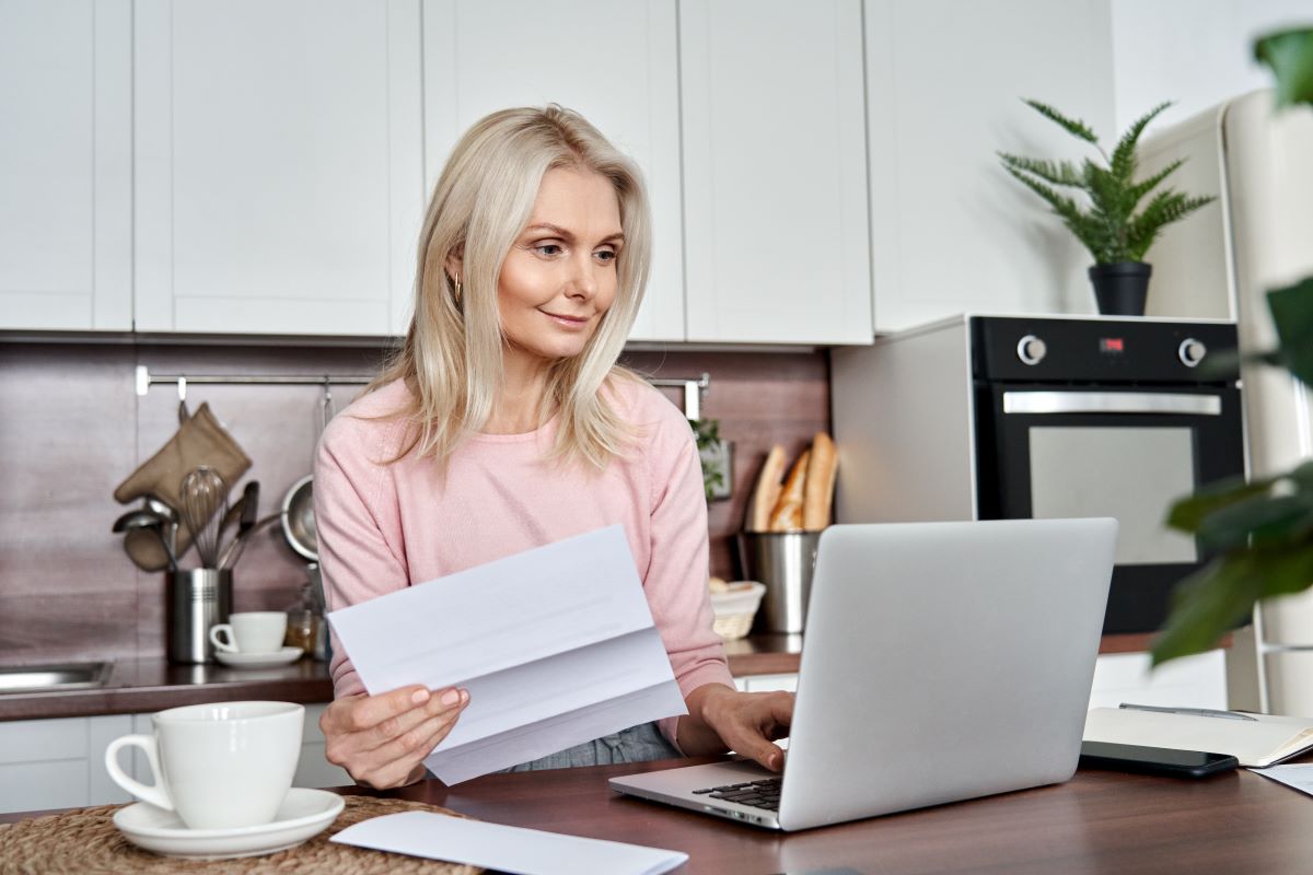 A middle-aged woman stands at her laptop in her kitchen, reviewing paperwork.